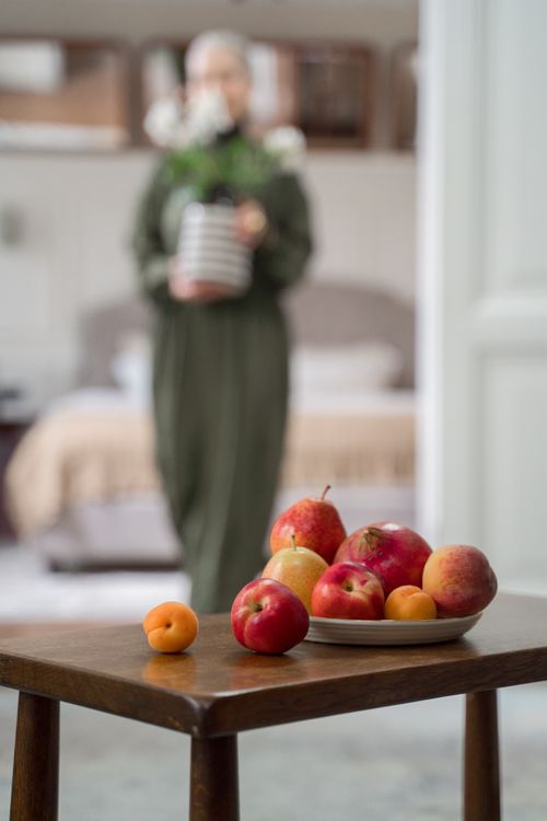 Free A plate of fresh fruits on a table with a blurred background of a woman holding flowers indoors. Stock Photo