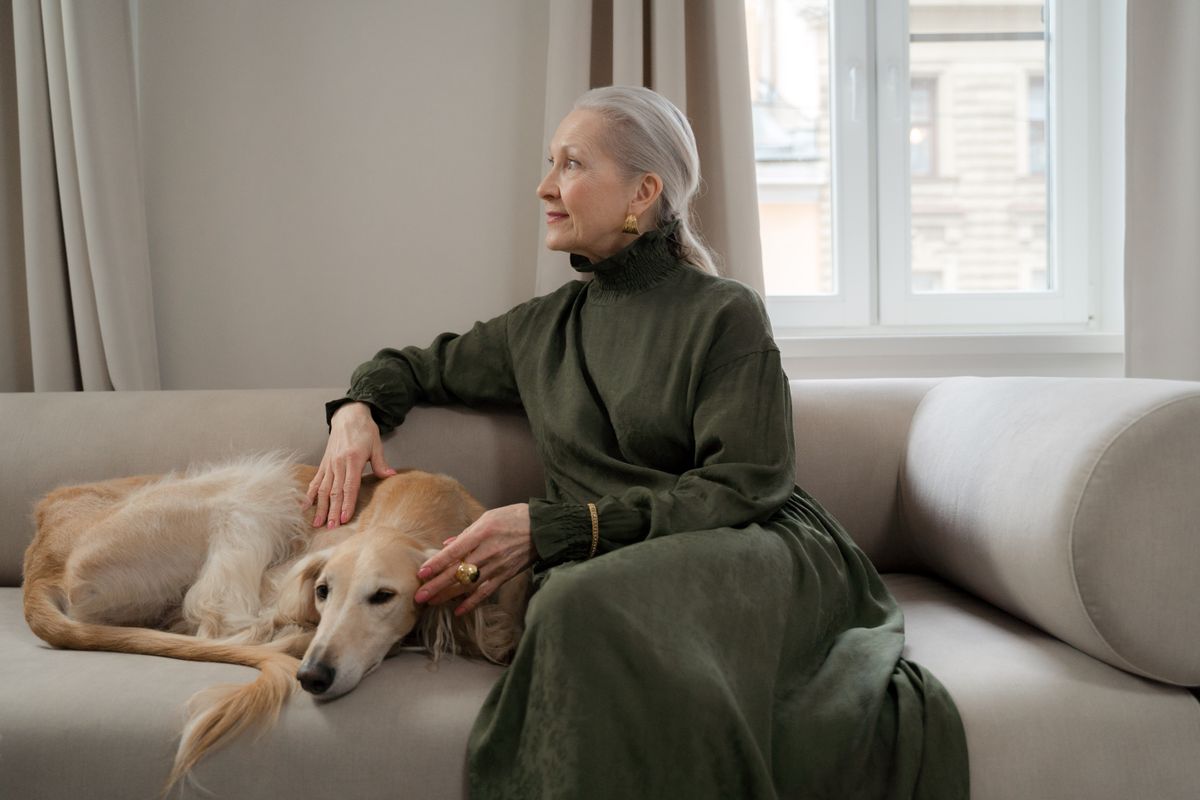 A greyhound rests beside a caregiver on a bright sofa where supplies can stay within reach.