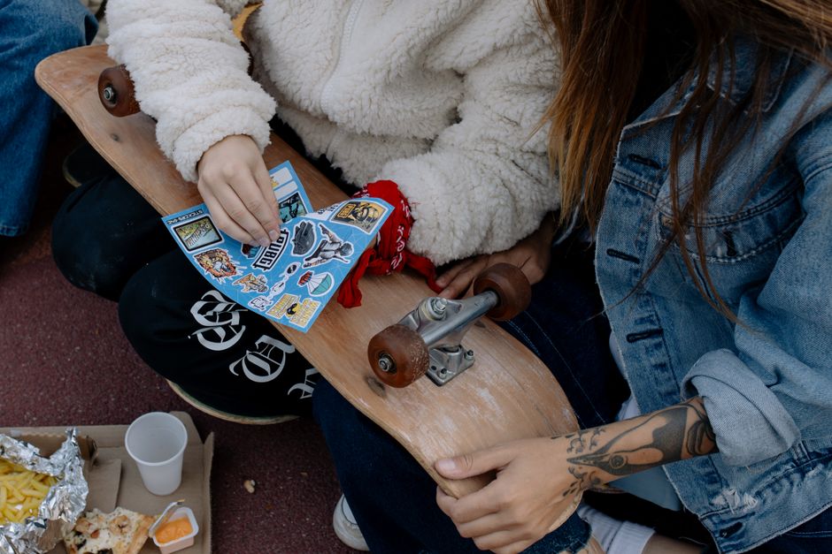 Two skaters customizing a skateboard with stickers while sitting outdoors, enjoying food.