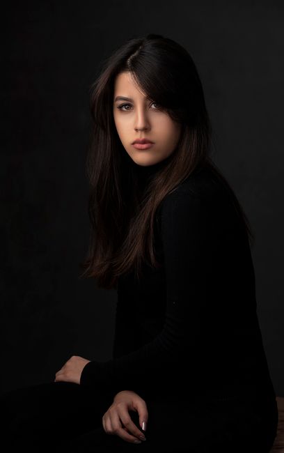 Elegant studio portrait of a young woman sitting against a dark background, gazing at the camera.