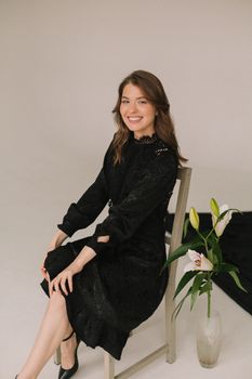 Smiling woman in a black dress sits beside lush flowers in a serene studio setting.