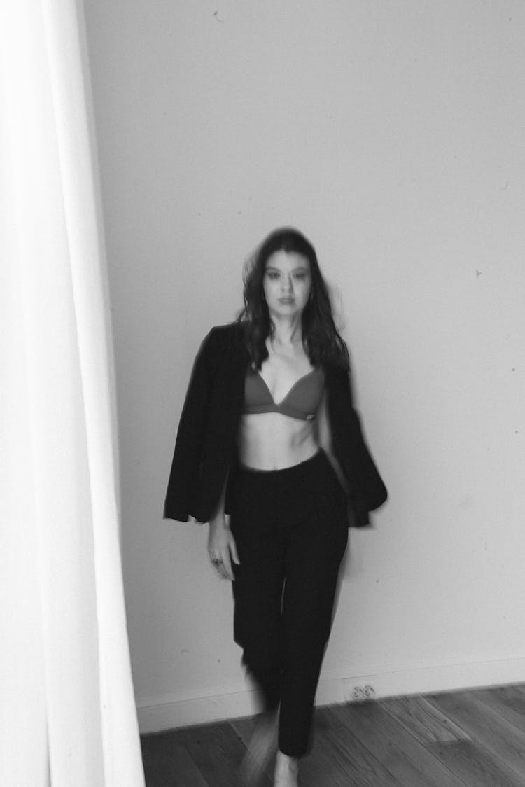 Grayscale Photo Of A Woman Standing With Black Blazer On Her Shoulder Wearing Bra And Black Pants 