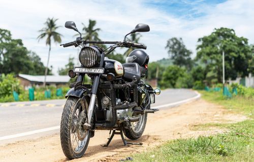 Free Black vintage motorcycle parked on a scenic rural road with lush greenery and clear blue sky. Stock Photo