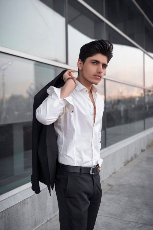 Free A fashionable young man poses with confidence beside a modern glass facade, wearing a black suit and white shirt in Tehran. Stock Photo