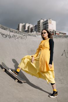 Stylish woman in yellow dress posing at skate park with skateboard. Urban fashion vibes.