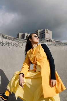 Fashionable young woman posing in a yellow dress at a skate park on a cloudy day.