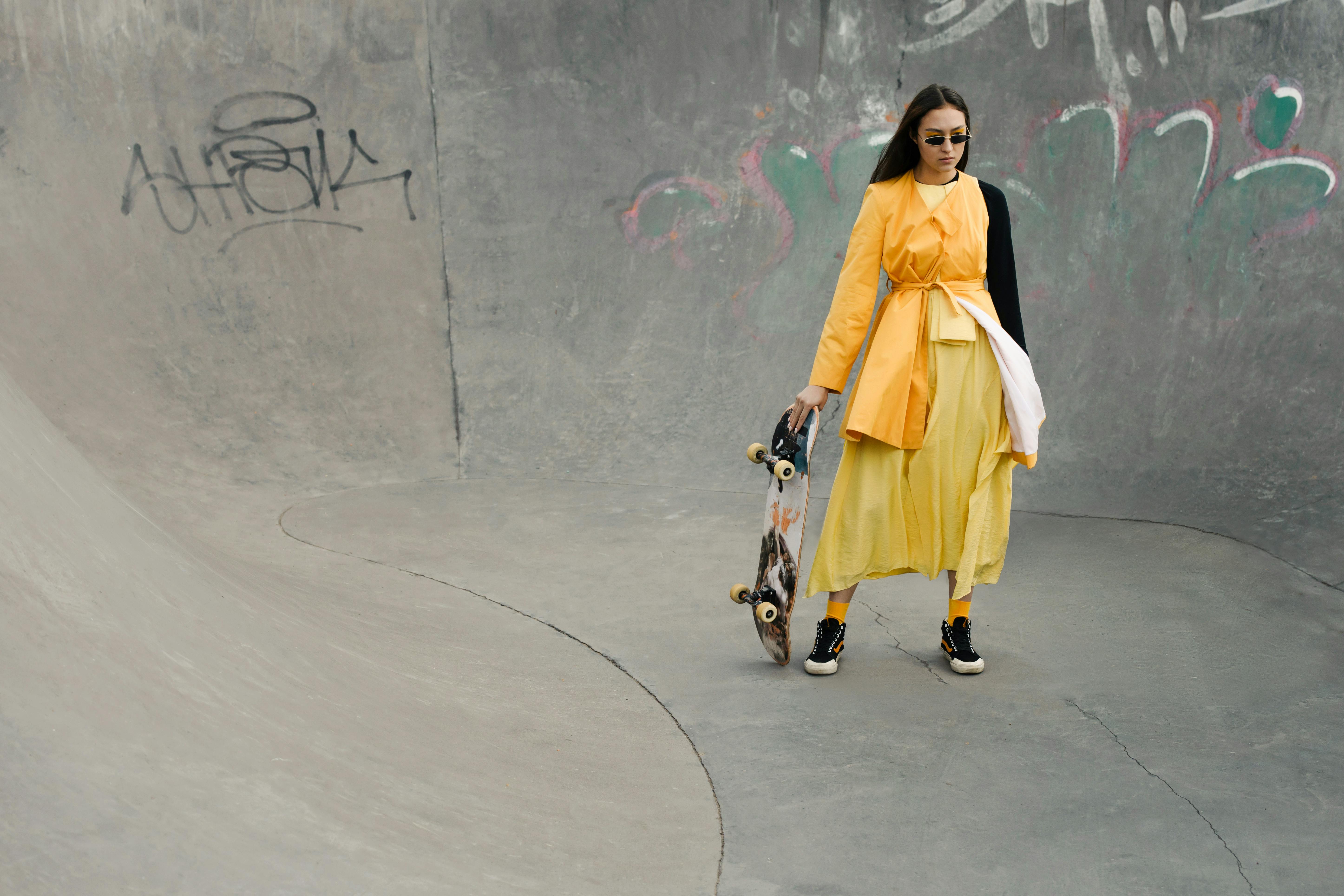 Chic woman in yellow dress holding a skateboard at a skatepark, embracing urban style.