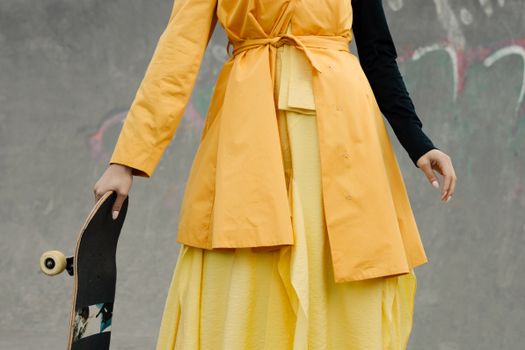 Stylish adult woman in a yellow dress holding a skateboard at a skate park, blending fashion and sport.