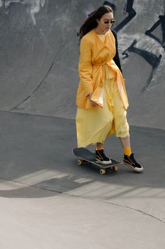 Fashionable Asian woman skateboarding in a vibrant yellow outfit at an outdoor skatepark.