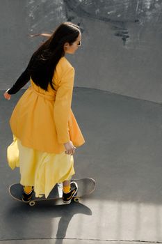 A stylish woman in yellow blazer and dress skateboarding at an urban park.