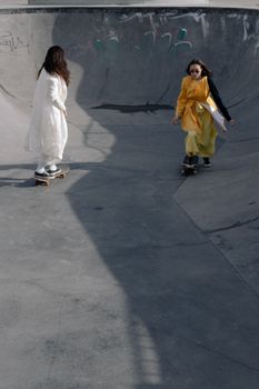 Two women in dresses skateboarding in a concrete skate park.