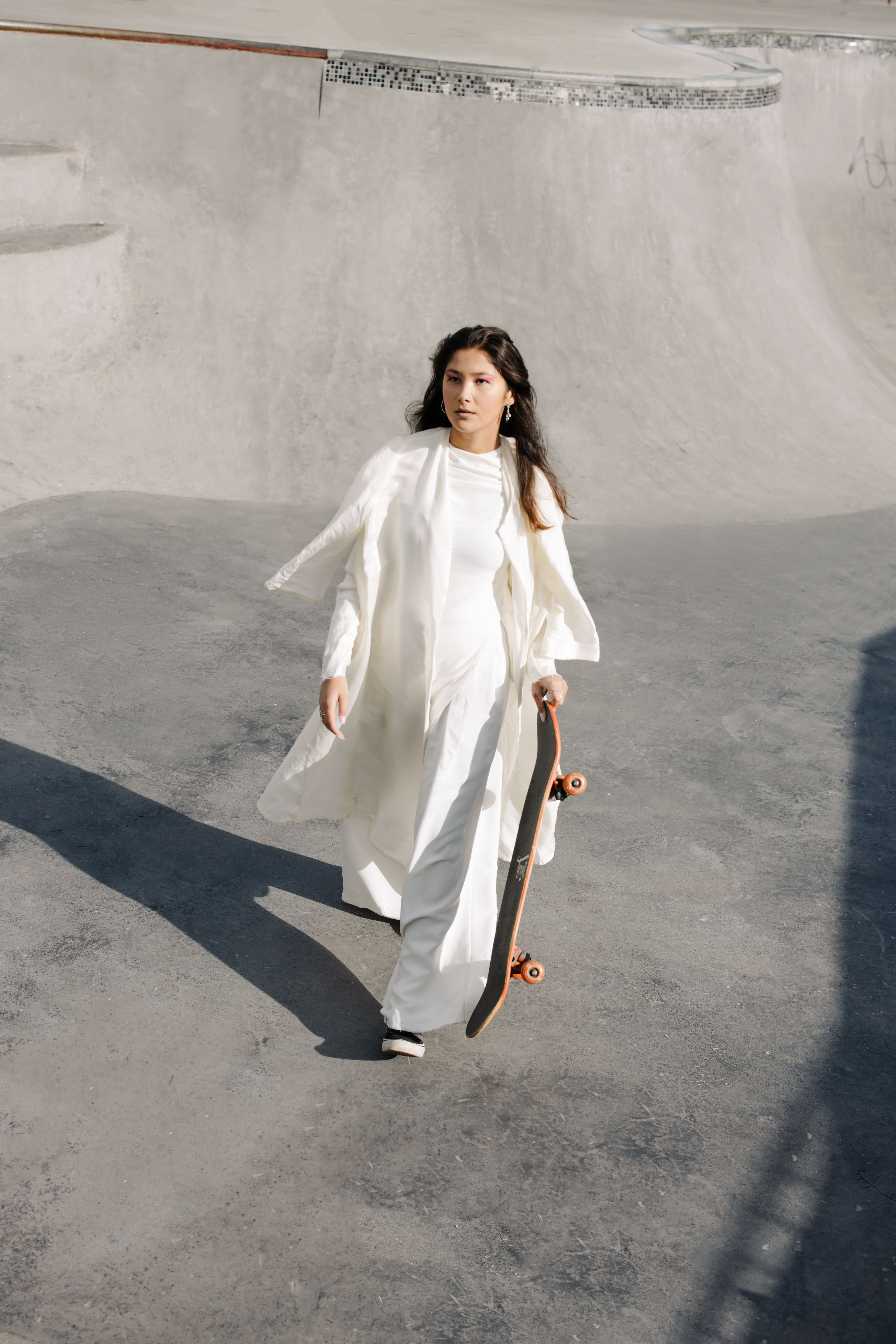 Elegant woman in white dress holding a skateboard in an urban skatepark, showcasing style and confidence.