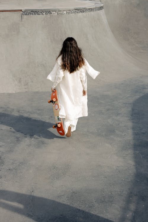 Person Walking in White Long Sleeve Dress Holding a Skateboard