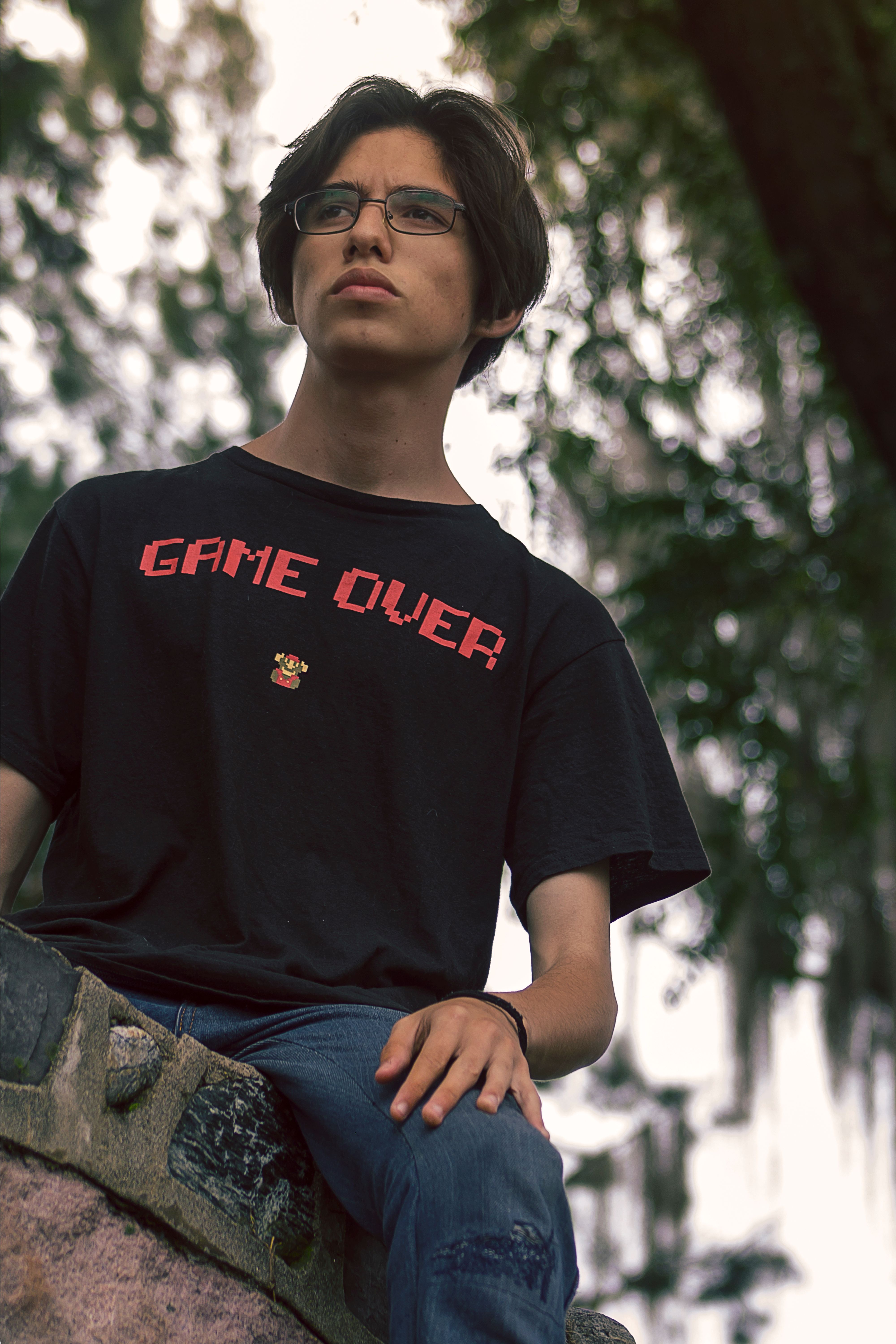 Young man sitting on a concrete ledge wearing a Game Over t-shirt in a natural outdoor setting.