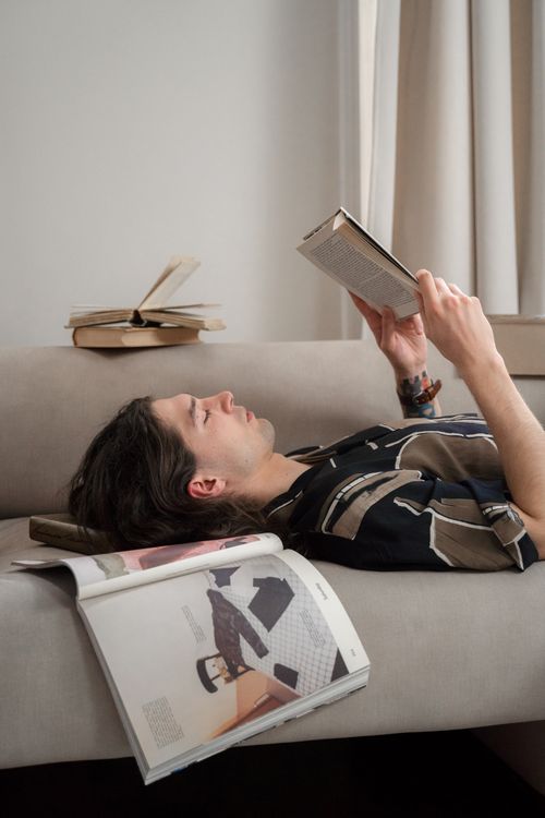 Free A relaxed young man lies on a sofa reading a book, surrounded by magazines in a cozy indoor setting. Stock Photo