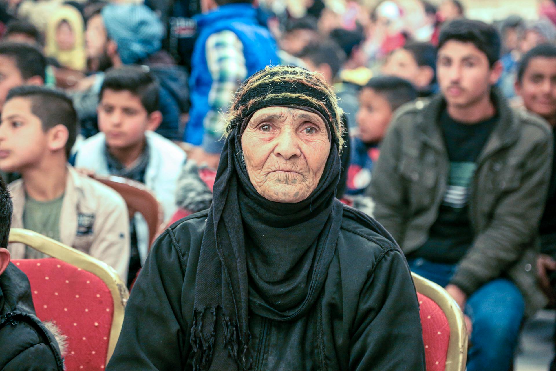 Elderly woman in black headscarf seated among crowd at community event in Lebanon.