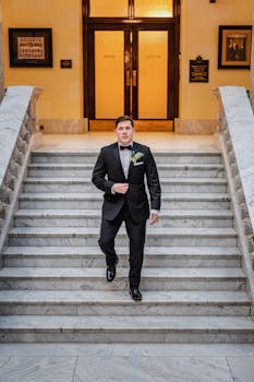 Groom in a tuxedo stands on marble stairs, ready for wedding ceremony.