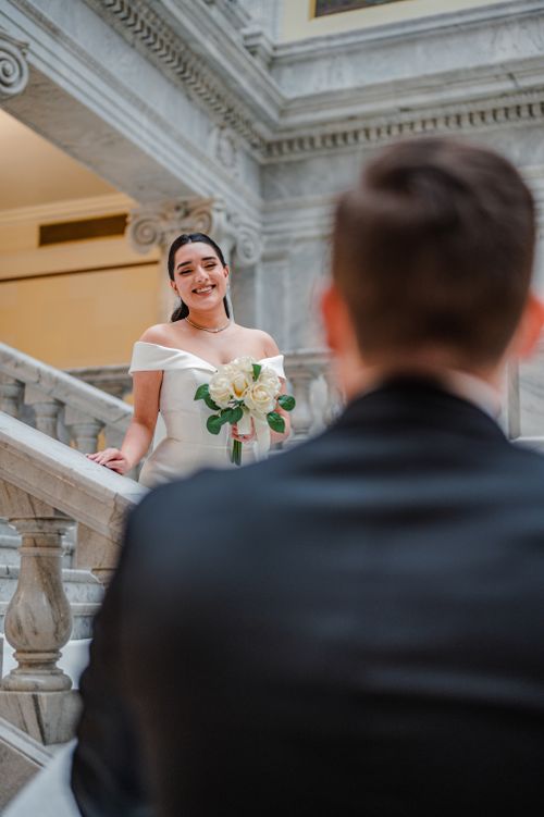 Free Bride in white dress smiles gracefully on marble staircase, bouquet in hand. Stock Photo