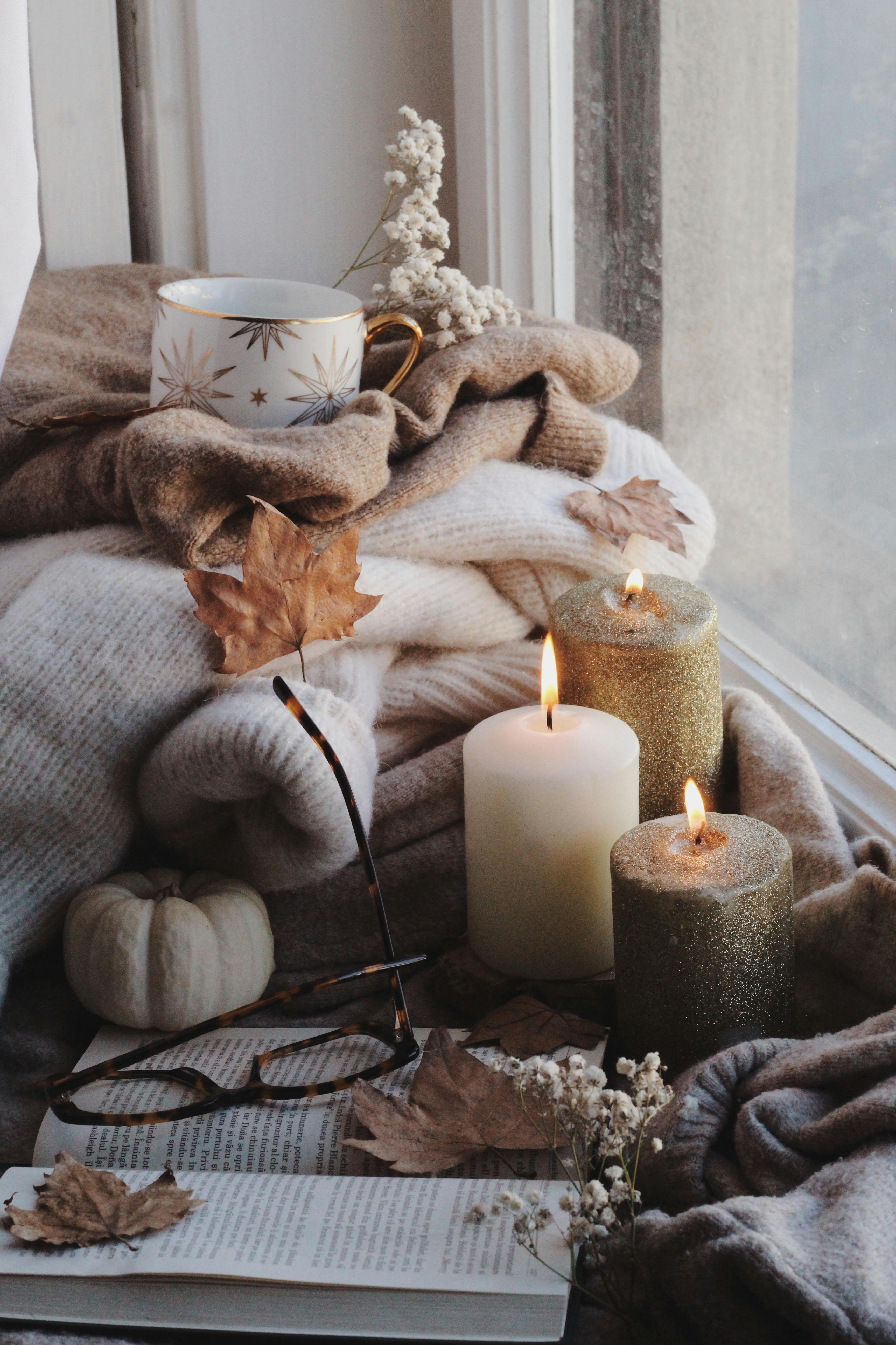 Comforting autumn setup by the window with candles, knitted sweaters, and a warm mug.