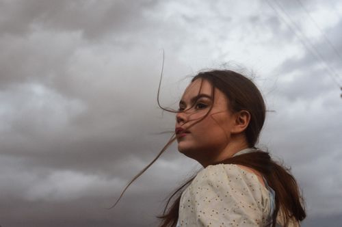 Free Profile view of a young woman with wind-swept hair looking at a cloudy sky, creating a dramatic outdoor portrait. Stock Photo