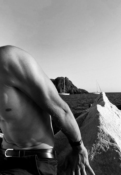 A Grayscale Photo of Topless Man Sitting on the Rock