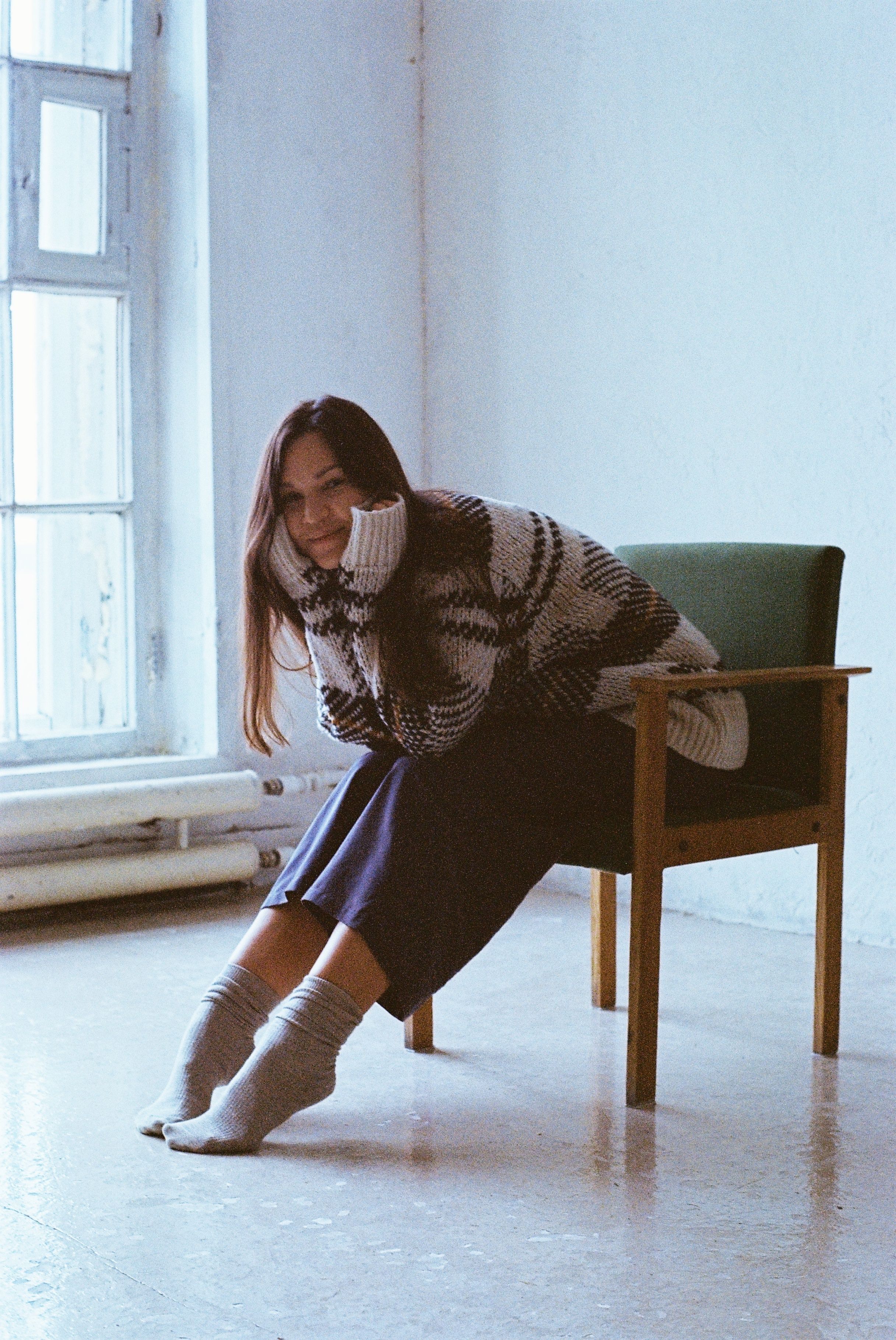 A woman in a cozy sweater sits and smiles indoors on a sunny day, Санкт-Петербург.