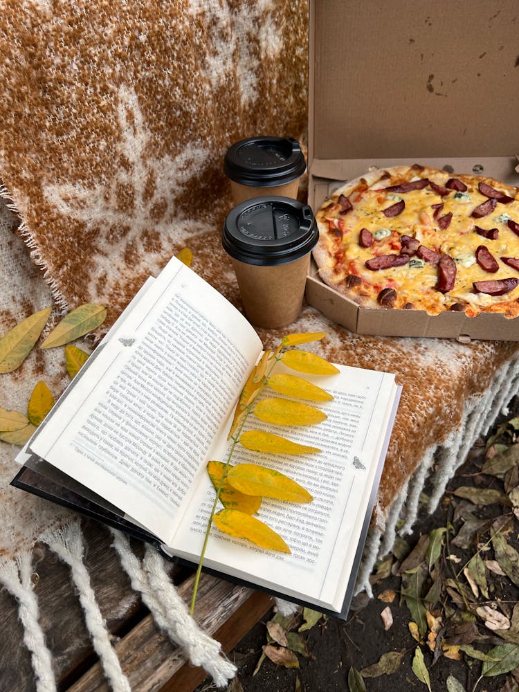 An Open Book With Leaves Near The Box Of Pizza And Coffee