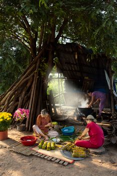 Women preparing traditional food outdoors by a rustic hut, under a large tree.