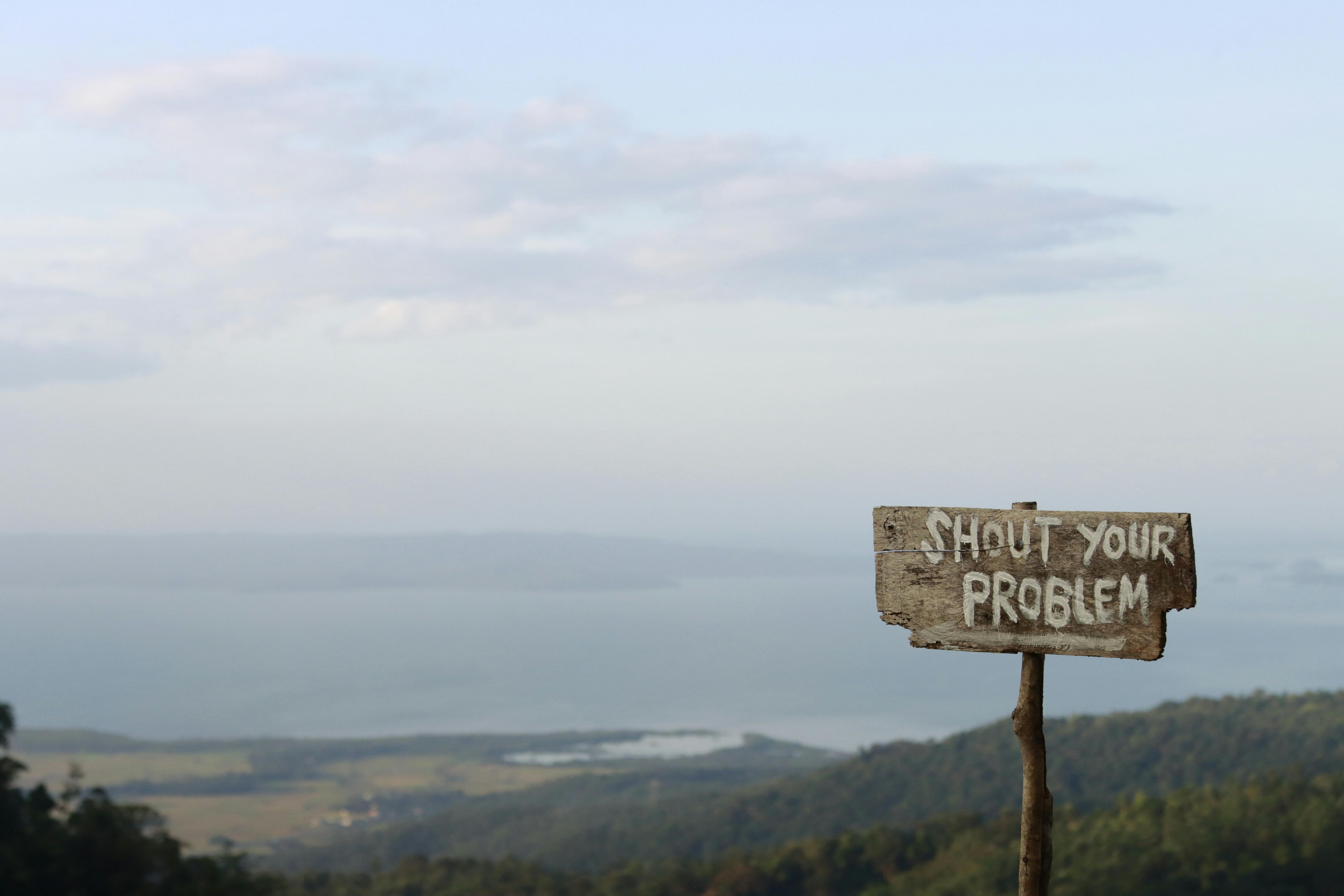 Wooden sign inscribed with 'Shout Your Problem' facing a beautiful mountain landscape under a blue sky.