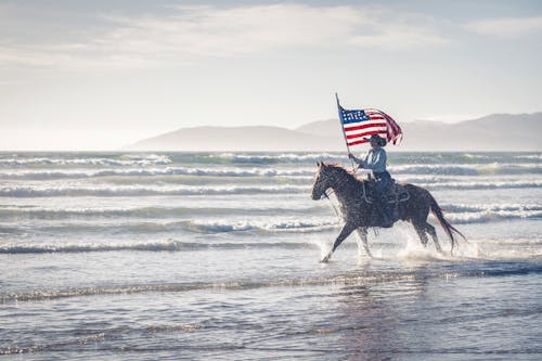 Woman Riding Horse Along Sea Holding American Flag