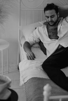 Man with beard and open shirt lying on bed in a relaxed pose in black and white photo.