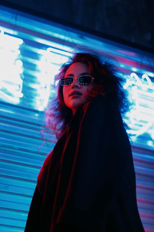 Free A fashionable woman wearing sunglasses stands illuminated by vibrant neon lights at night. Stock Photo