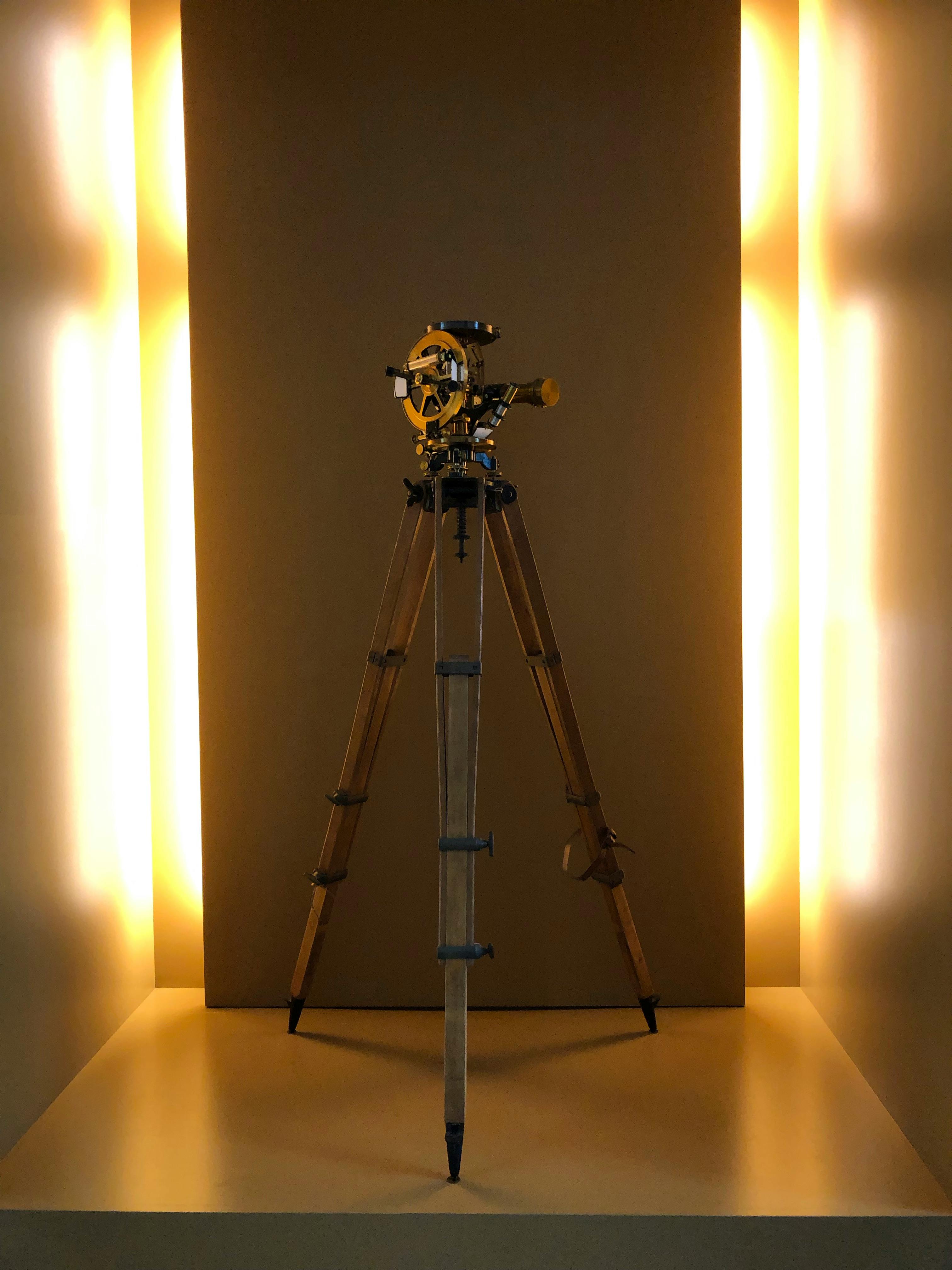A vintage theodolite stands on a wooden tripod, beautifully lit in a modern setting.