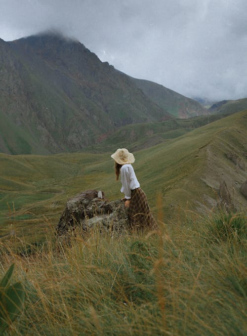 A Woman in White Long Sleeves Standing on Green Grass Field Near the Mountain