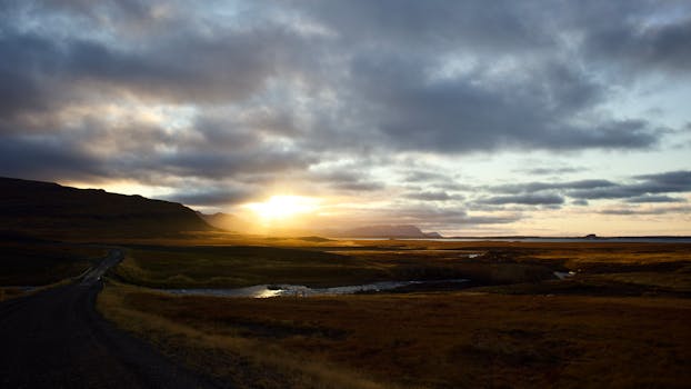 Captivating sunrise over an Icelandic landscape with road, hills, and river under a dramatic sky.