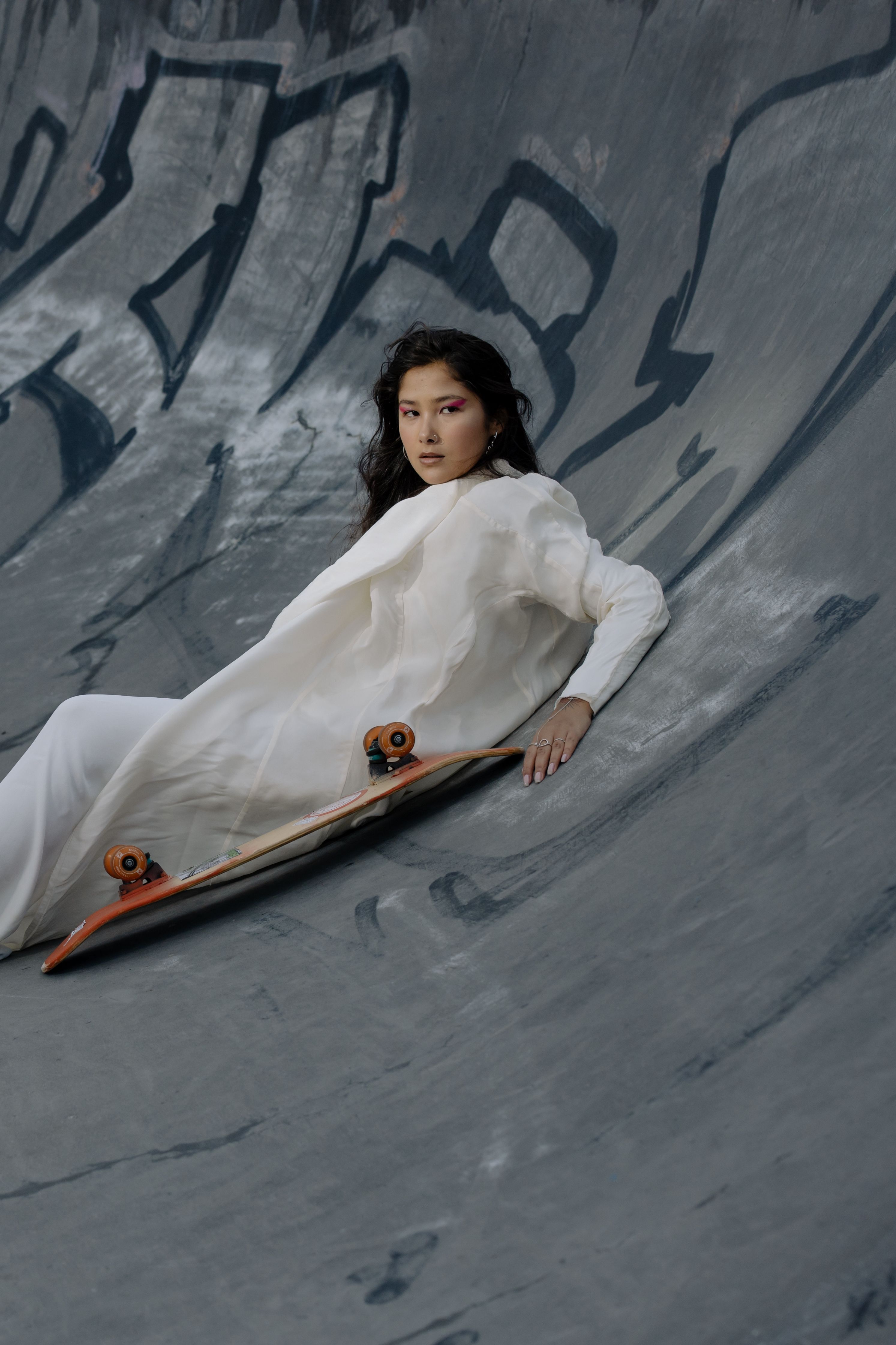 A woman in a white dress poses creatively in a concrete skate park, blending fashion and skate culture.
