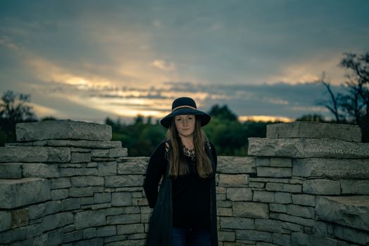 A woman with long hair and a hat stands by a stone wall during an overcast dusk.