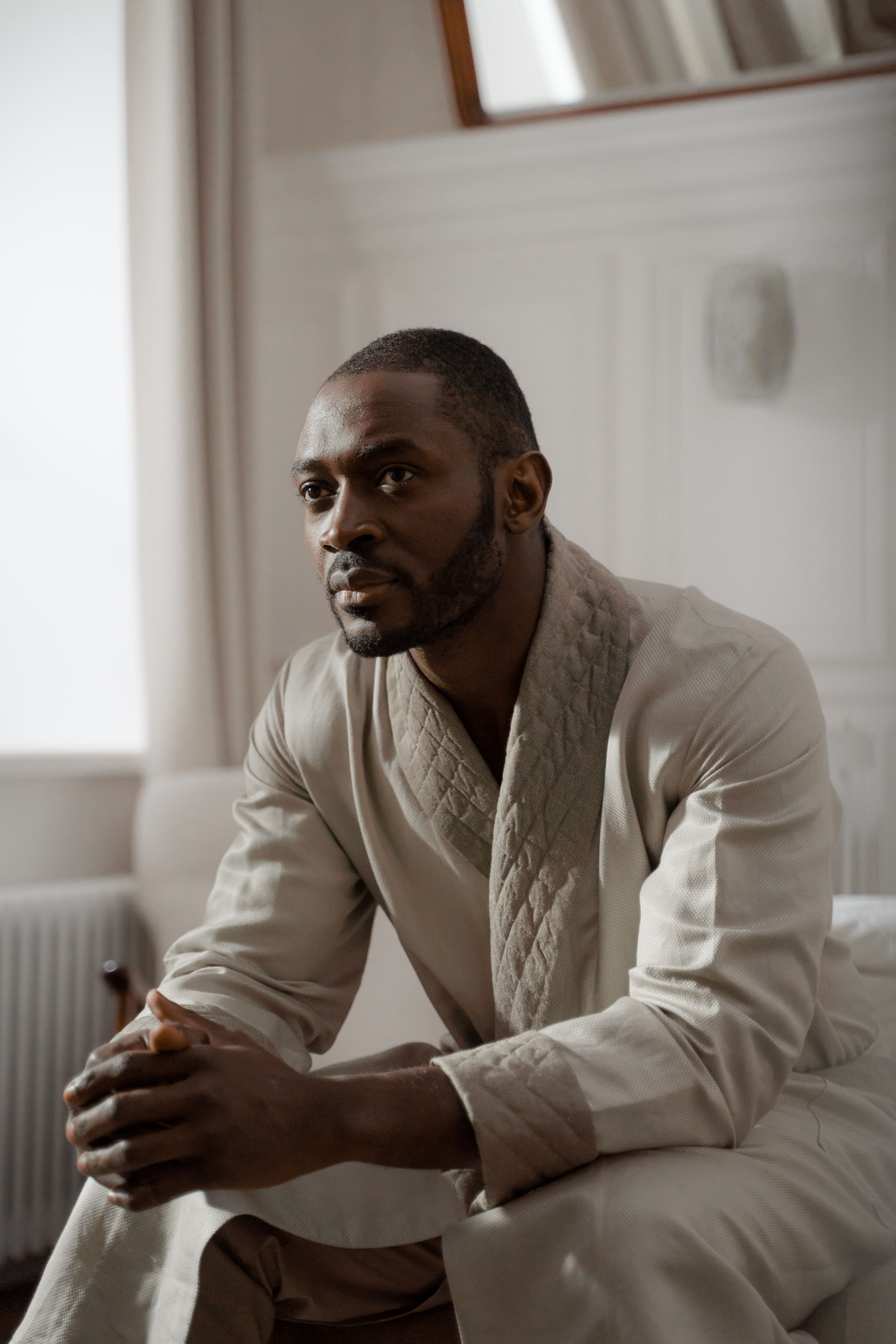 A man in a bathrobe sits thoughtfully in a bright, cozy living room, bathed in natural morning light.