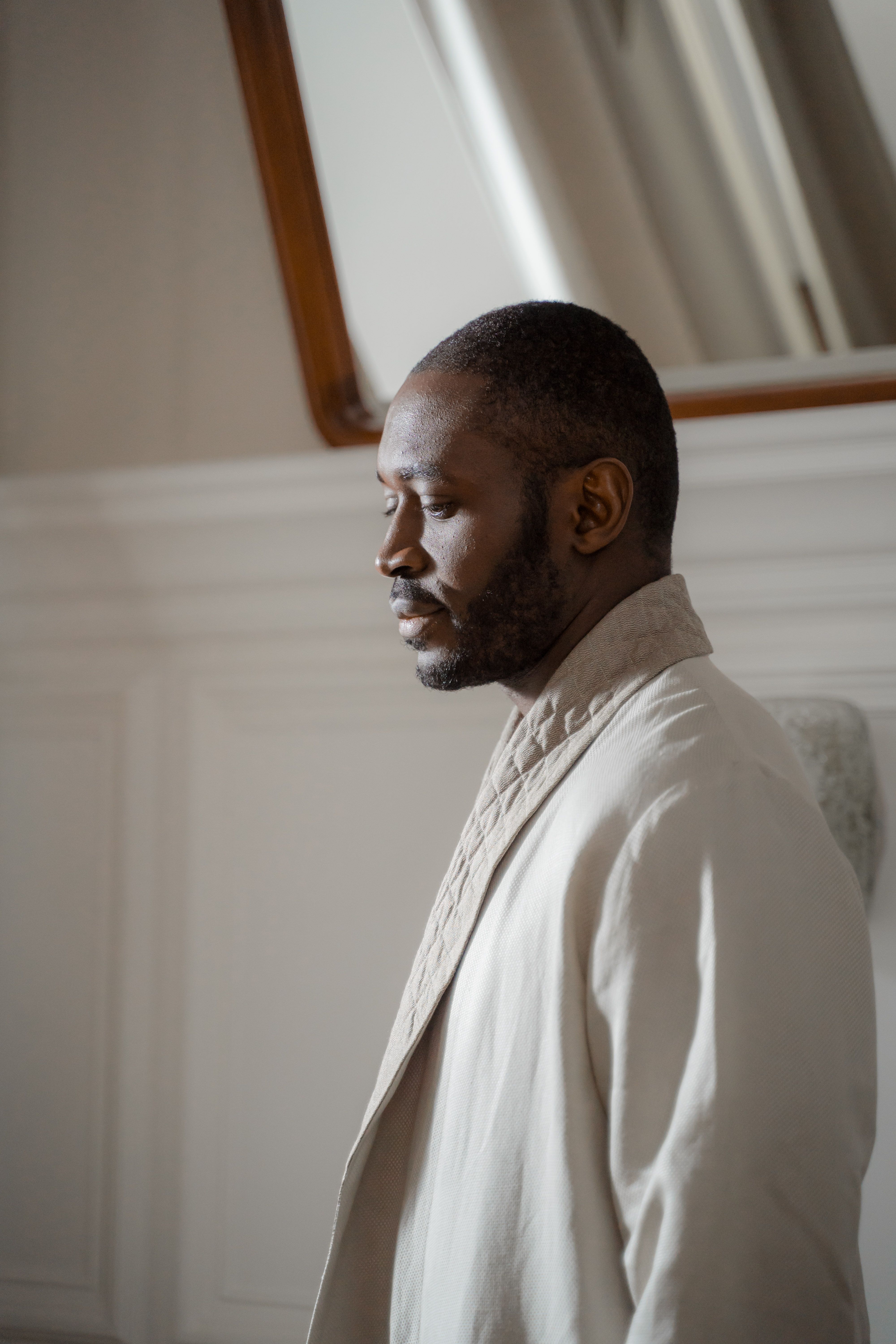 A contemplative man in a light bathrobe stands indoors, captured in profile with natural light.