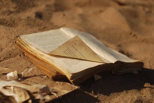 An open, aged book resting on sandy ground under warm daylight, highlighting its vintage charm.