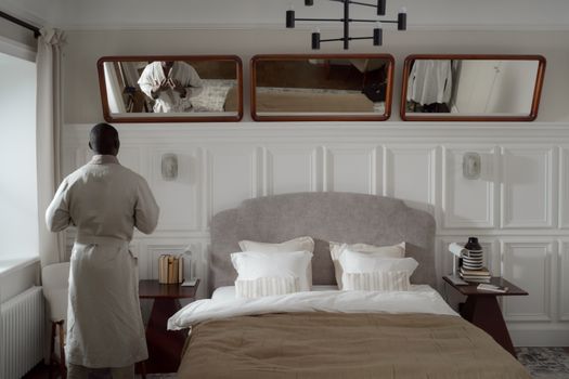 Adult man in bathrobe adjusting clothing in a stylish bedroom setting with mirrors.