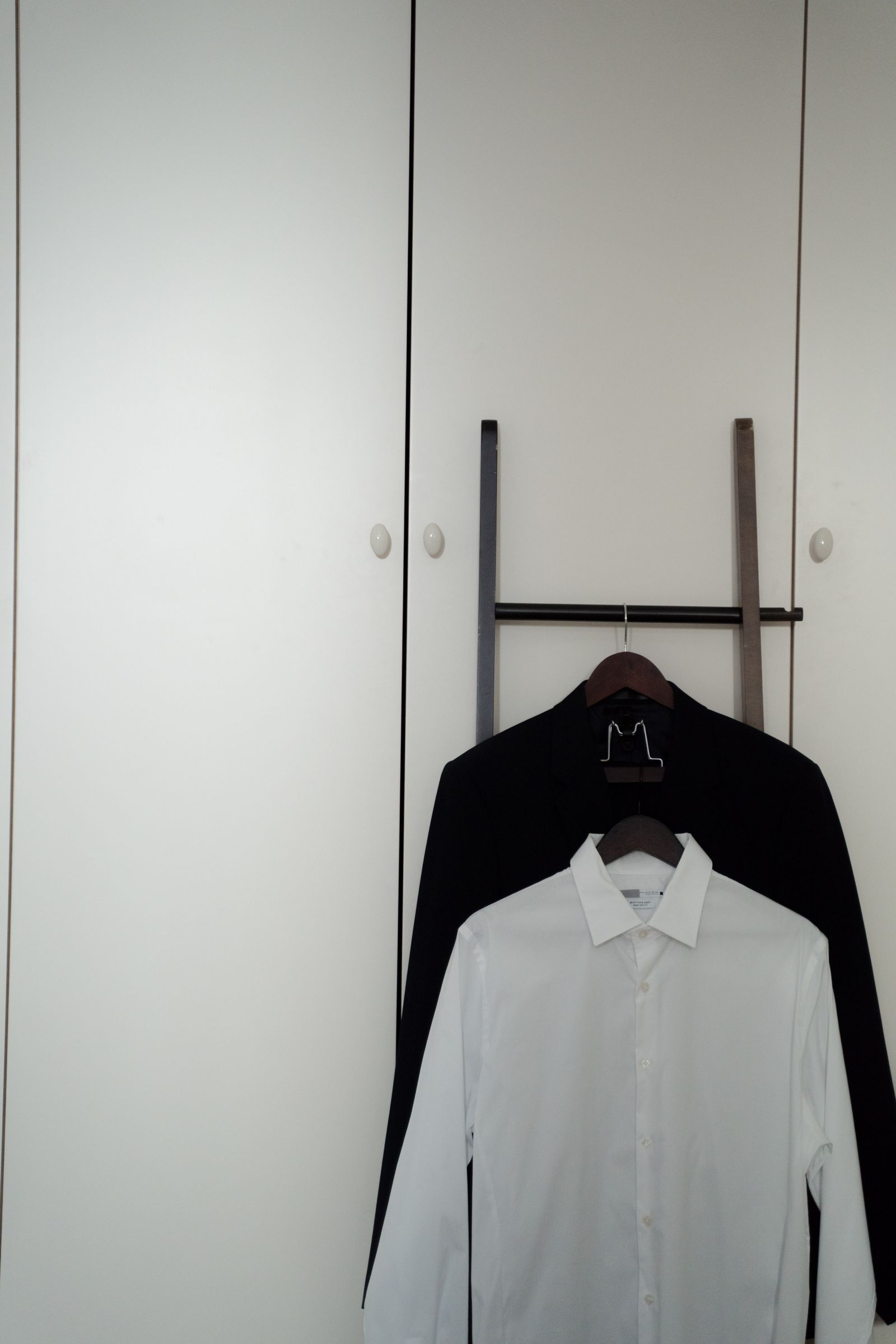 A black shirt and a white shirt hung on a ladder rail against white wardrobe doors