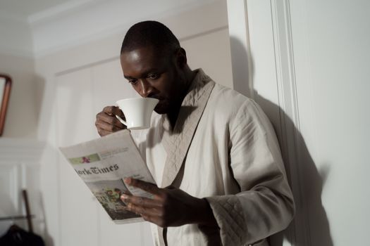 Man in a bathrobe enjoys coffee and reads a newspaper indoors.