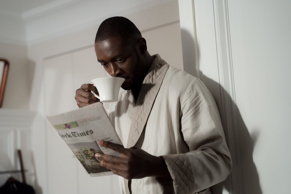 Man in a bathrobe enjoys coffee and reads a newspaper indoors.
