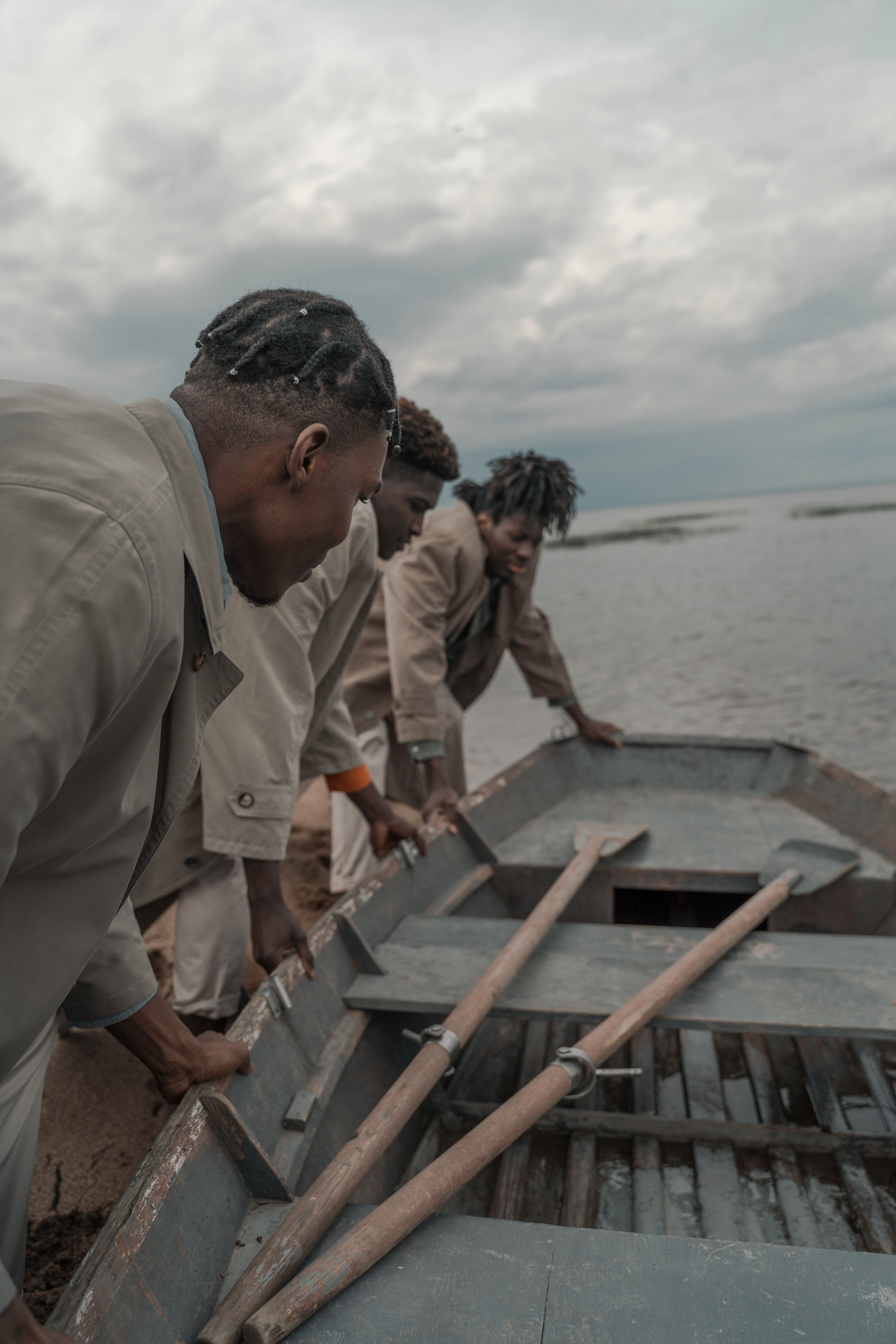 Three men with braided hair prepare a wooden boat by the sea under an overcast sky.