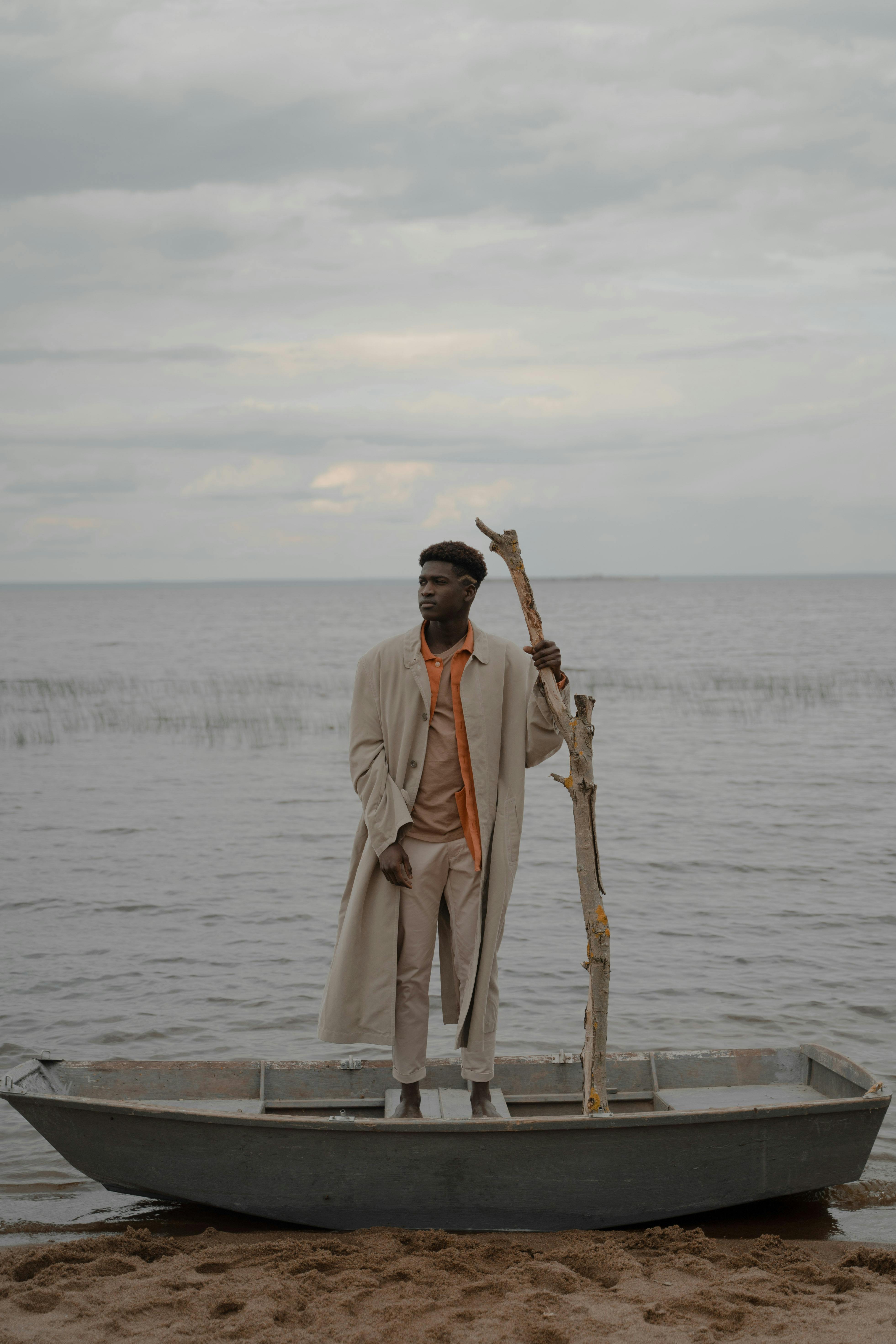A fashionable man stands confidently in a boat on a calm beach, wearing a coat and holding a branch.