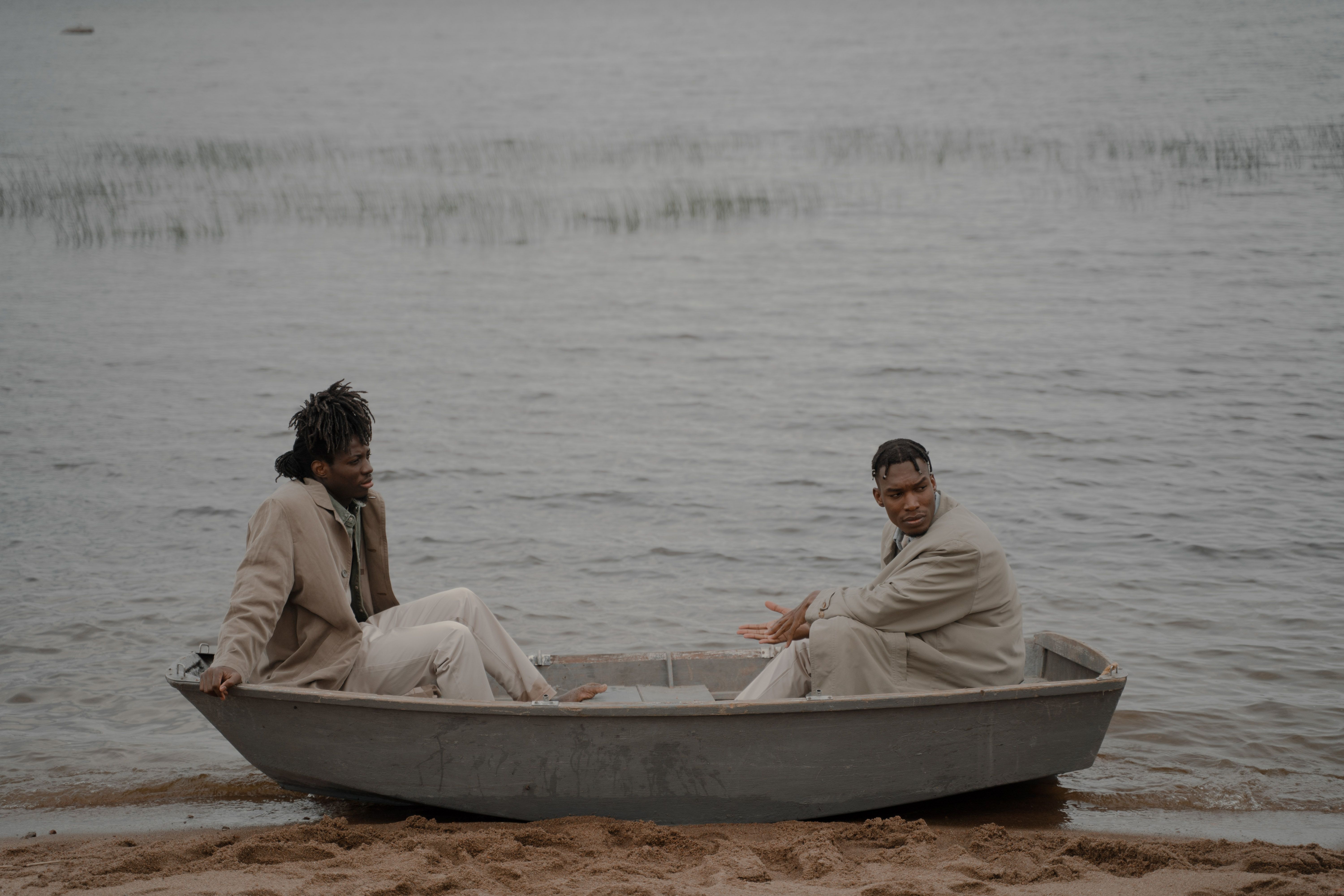 Two men in coats sitting in a boat by the beach with calm sea backdrop.