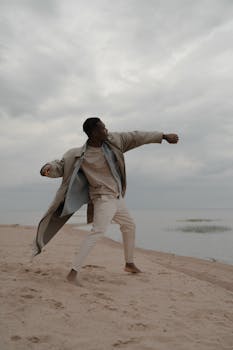 A stylish man dances barefoot on a sandy beach under a moody twilight sky, exuding freedom.