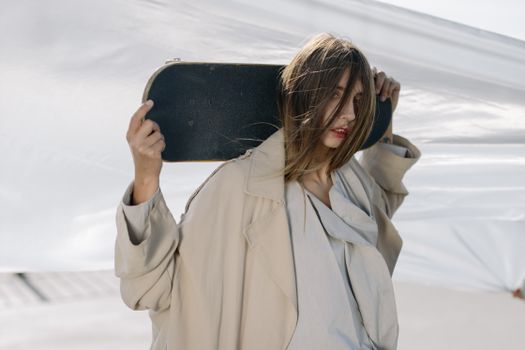 Caucasian woman in a trench coat holding a skateboard outdoors. Fashionable and modern style.