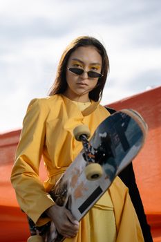 Fashionable young woman holding a skateboard with a confident look, wearing a yellow trench coat and sunglasses.